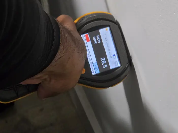 Technician using a moisture meter against a wall with a reading of 26.5